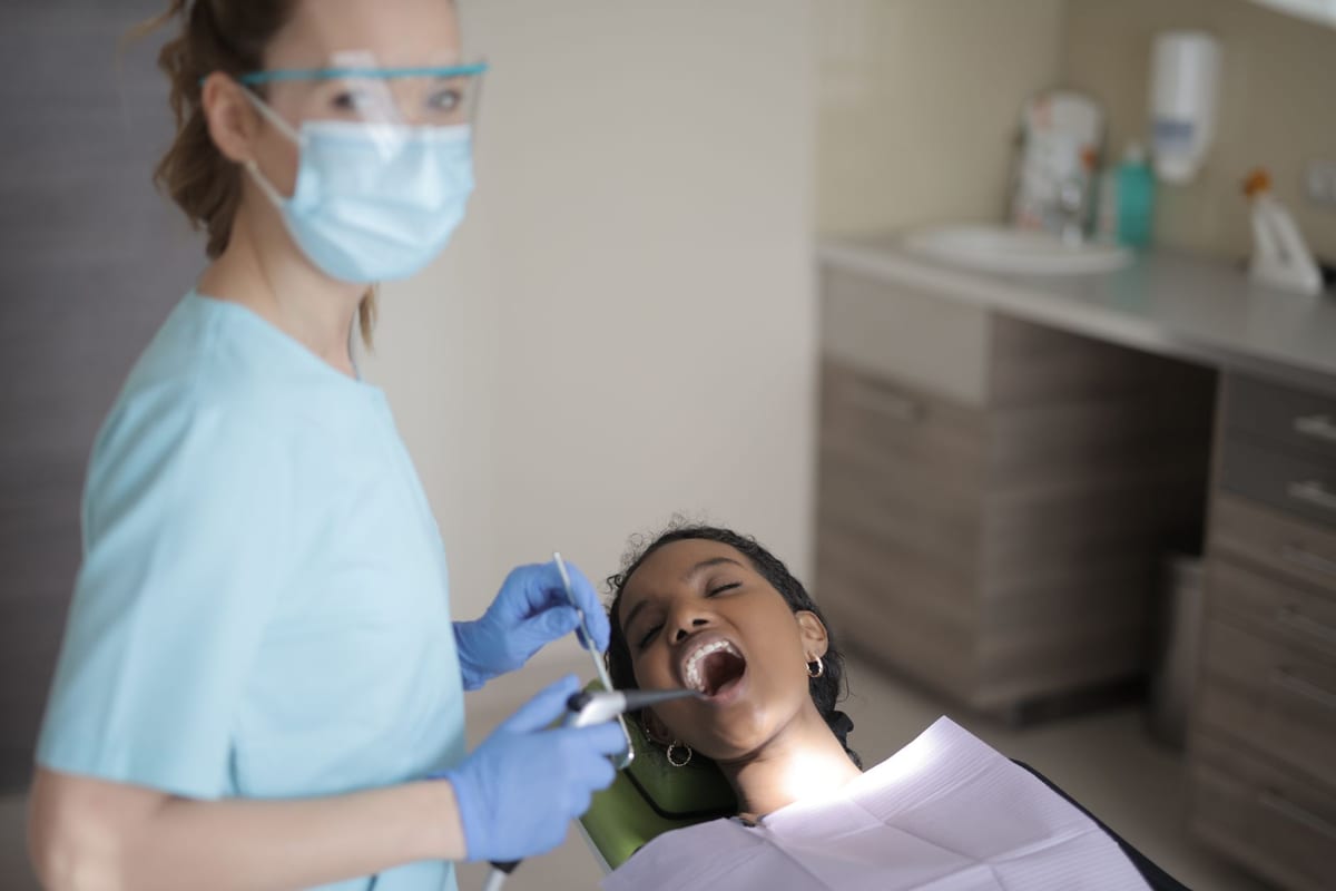 A hygienist in a mask and eye protection cleaning a seated patient's teeth